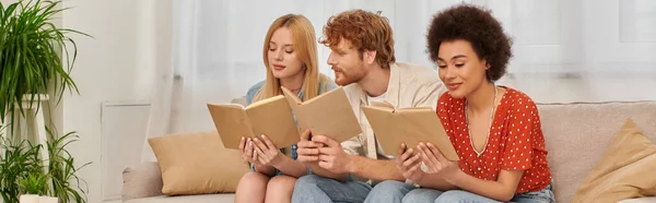 Alternative relationships, polygamy concept, intelligent multicultural women reading books with redhead boyfriend in living room, modern family, hobby and leisure, freedom in relationship, banner — Stock Photo