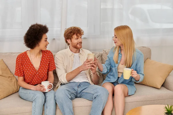 Família poliamorosa, diversidade de relacionamento, amantes da poligamia feliz sentados no sofá com xícaras de café, homem interracial e mulheres na sala de estar, bissexuais e pessoas poliamor — Fotografia de Stock