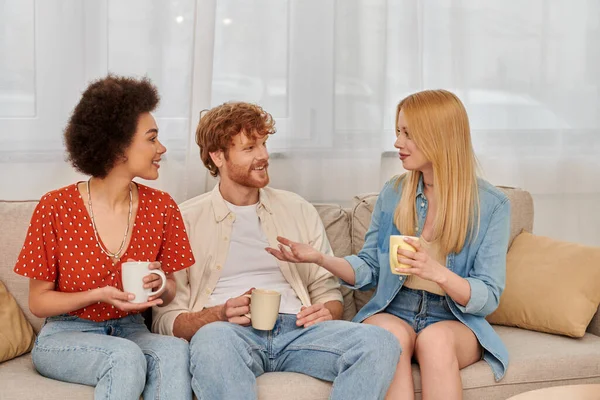Polyamouröse Familie, Beziehungsvielfalt, fröhliche Polygamie-Liebhaber, die auf der Couch sitzen und bei Tassen Kaffee plaudern, gemischtrassige Männer und Frauen im Wohnzimmer, bisexuelle und polyamory Menschen — Stockfoto