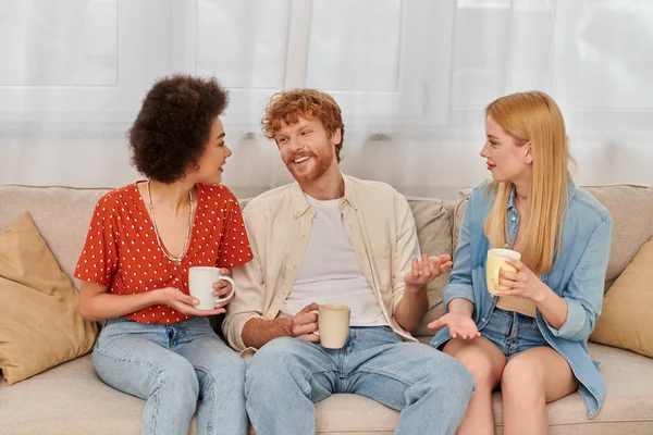 Concetto poligamo, diversità di relazione, felici amanti della poligamia seduti sul divano e con in mano tazze di caffè, uomo e donna interrazziale in salotto, bisessuale e poligamia — Foto stock