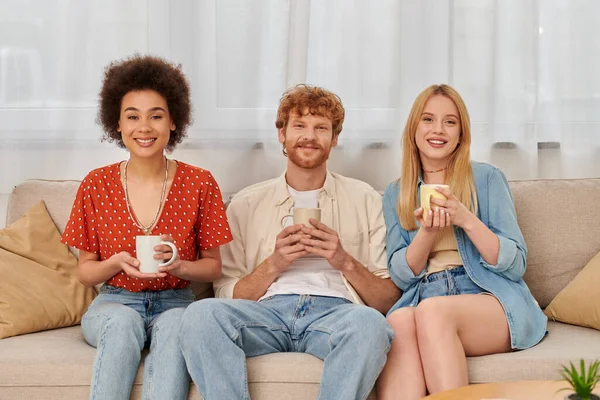 Polyamorous concept, relationship diversity, happy polygamy lovers sitting on couch and holding cups of coffee, looking at camera, interracial man and women in living room, bisexual and polygamy — Stock Photo