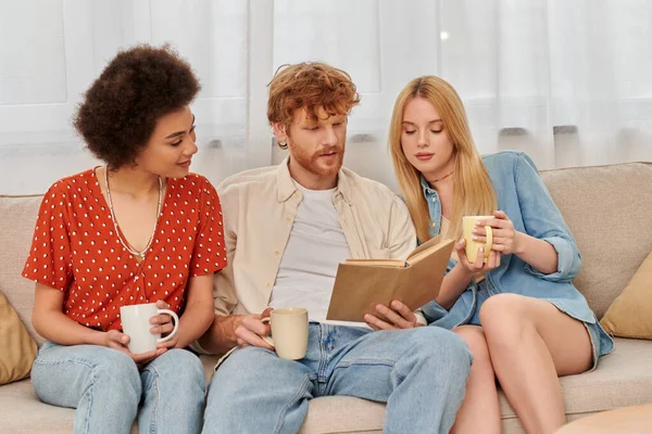 Alternative relationships, polygamy, intelligent redhead man reading book to multicultural women in living room, modern family, hobby and leisure, polyamorous family, cups of coffee — Stock Photo