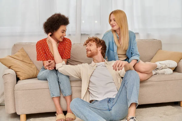 Love triangle concept, diversity in relationships and culture, non monogamy, happy redhead man sitting near multicultural women in living room, lovers, modern family, acceptance, open relationship — Stock Photo