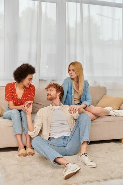Love triangle concept, diversity in relationships and culture, non monogamy, cheerful redhead man sitting near beautiful multicultural women in living room, lovers, modern family, open relationship — Stock Photo