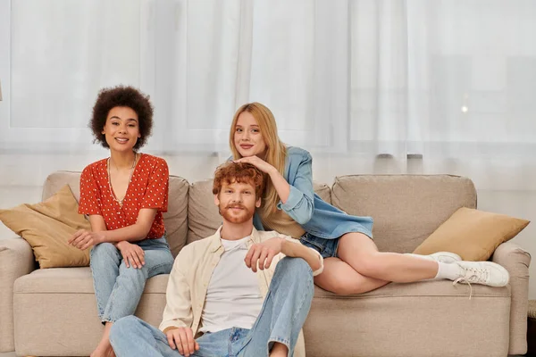 Concepto de poligamia, diversidad en las relaciones y la cultura, no monogamia, hombre pelirrojo feliz y mujeres multiculturales mirando a la cámara en la sala de estar, amantes, aceptación, relación abierta, bandera - foto de stock