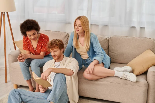 Open relationships, polygamy concept, intelligent man reading book to beautiful multicultural lovers in living room, modern family, hobby and leisure, polyamorous family, man and women — Stock Photo