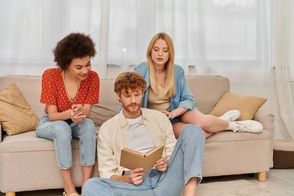 Alternative relationships, polygamy concept, intelligent man reading book to beautiful multicultural lovers in living room, modern family, hobby and leisure, polyamorous family, man and women — Stock Photo