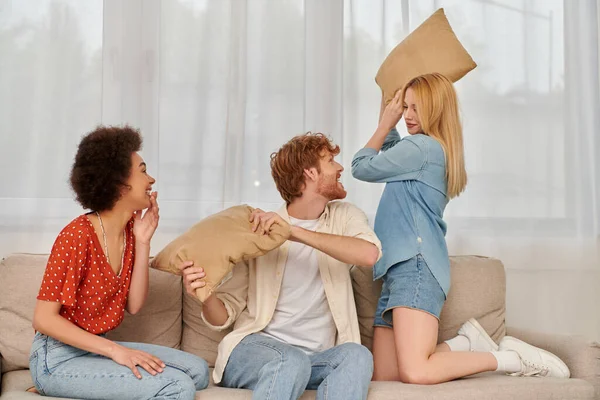 Polyamorous concept, group relationship, cheerful multicultural women and bearded man fighting with pillows, open relationship, diversity and bonding, non monogamy, three people — Stock Photo