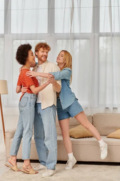 Família alternativa, mulheres multiculturais felizes abraçando o homem ruivo na sala de estar, diversidade cultural, liberdade nos relacionamentos, triângulo amoroso, bissexuais e pessoas de espírito livre — Fotografia de Stock