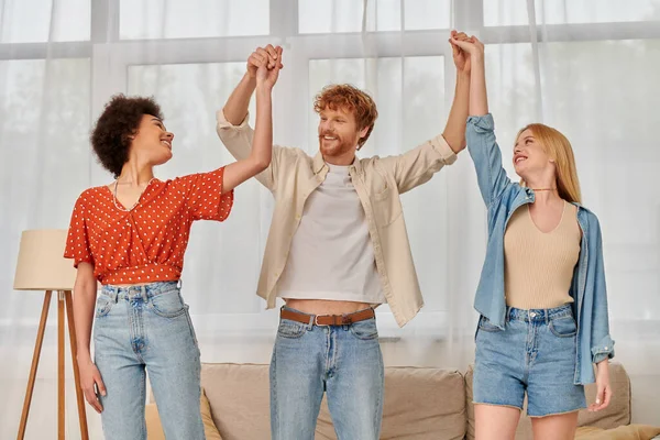 Polygamy, three people holding and raising hands while standing in living room, alternative family, acceptance and understanding in relationship, bonding and love, multiracial lovers — Stock Photo