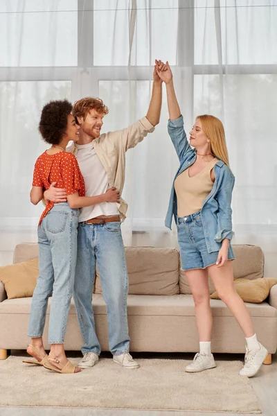 Polygamy concept, lovers holding and raising hands while standing in living room, alternative family, acceptance and understanding in relationship, bonding and love, multiracial lovers — Stock Photo