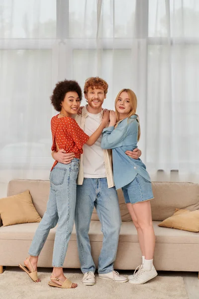 Polygamy and love concept, three adults, happy redhead man hugging with multicultural women, threesome, cultural diversity, acceptance, bonding and love, multiracial lovers in living room — Stock Photo