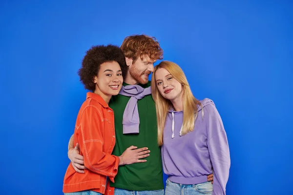Polyamory concept, cheerful multicultural women looking at camera and hugging with redhead man on blue background, studio photography, cultural diversity, polygamy, young lovers in casual attire — Stock Photo