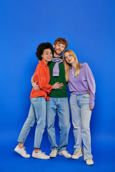 Alternative relationships, polyamory, three people, young redhead man and beautiful multiracial women on blue background, studio shot, vibrant colors, casual clothes, stylish attire, modern family — Stock Photo