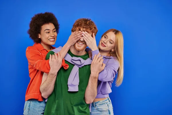 Polyamory, three people, beautiful multiracial women covering eyes of young redhead man on blue background, studio shot, vibrant colors, casual attire, modern family, alternative relationships, — Stock Photo