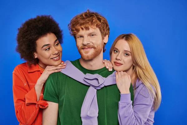 Alternative relationships, polyamory, portrait of three people, young redhead man and beautiful multiracial women on blue background, studio shot, vibrant clothes, stylish attire, modern family — Stock Photo