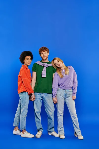 Alternative family, polygamy, portrait of three people, young redhead man and beautiful multiracial women on blue background, studio shot, vibrant clothes, polyamory, modern relationships — Stock Photo