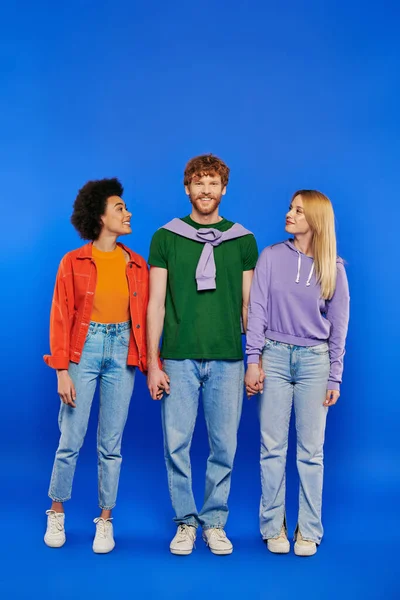 Polyamory three people, young man and women holding hands on blue background, studio shot, vibrant clothes, modern love triangle, alternative relationships, polygamy lovers, happiness — Stock Photo