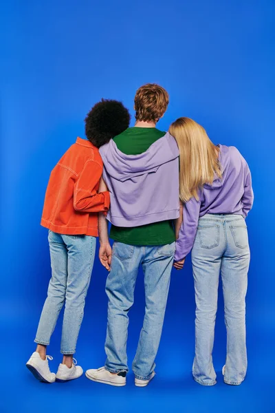 Poligamia, visão traseira de poliamor três pessoas, homem jovem e mulheres de mãos dadas sobre fundo azul, tiro de estúdio, moda jeans, triângulo amoroso, ligação, comprimento total — Fotografia de Stock