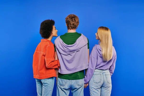 Polygamy, back view of polyamory three people, young man and multicultural women holding hands on blue background, studio shot, denim fashion, love triangle, bonding and love — Stock Photo