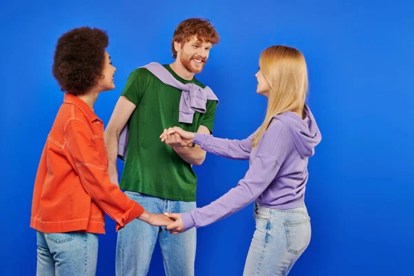Polyamorous relationship concept, polygamy lovers, young man and multicultural women holding hands on blue background, studio shot, denim fashion, love triangle, bonding and acceptance — Stock Photo