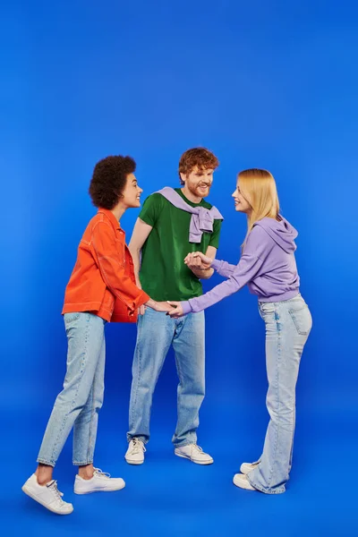 Concepto de familia poliamorosa, amantes de la poligamia, hombre joven y mujeres bisexuales multiculturales cogidas de la mano sobre fondo azul, plano de estudio, moda vaquera, triángulo amoroso, vinculación y aceptación - foto de stock