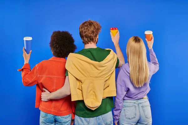Love triangle, polyamory lovers, back view of young redhead man and multiracial women holding coffee to go on blue background, studio shot,  takeaway drink, vibrant colors, modern family — Stock Photo
