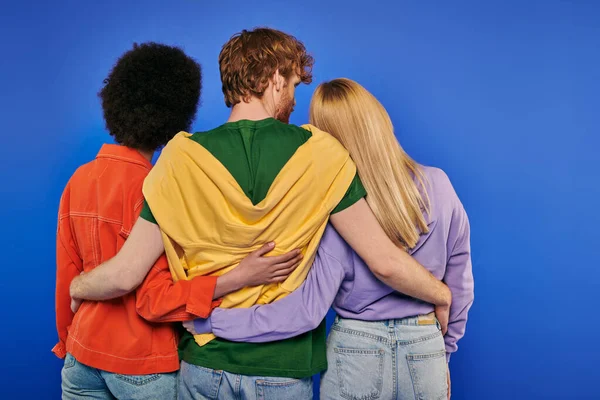 Love triangle, polyamory lovers, back view of young redhead man hugging multiracial women on blue background, studio shot, vibrant colors, casual clothes, stylish attire, modern family — Stock Photo