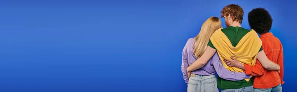 Amore triangolo, poliamore, tre persone, vista posteriore di uomo rossa abbracciando donne multirazziali su sfondo blu, riprese in studio, colori vivaci, abiti casual, abbigliamento elegante, famiglia moderna, banner — Foto stock