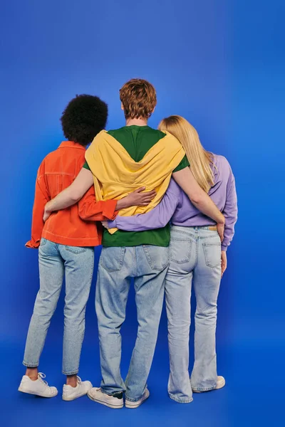 Любовний трикутник, поліаморія, три людини, вид ззаду молодого рудого чоловіка, що обіймає багаторасових жінок на синьому фоні, студійний знімок, яскраві кольори, повсякденний одяг, стильний одяг, сучасна сім'я — стокове фото