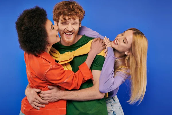 Alternative relationships, polyamory lovers, cheerful multiethnic women kissing and hugging young redhead man on blue background, studio shot, vibrant colors, modern family, bonding and acceptance — Stock Photo
