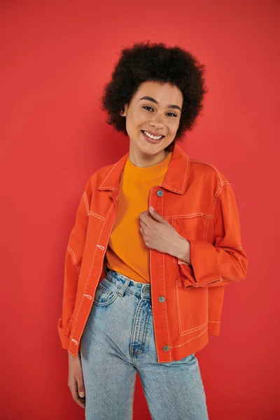 Happy young african american woman posing in stylish and vibrant outfit on coral background, smiling and looking at camera, fashion statement, African beauty, natural curly hair, trend — Stock Photo