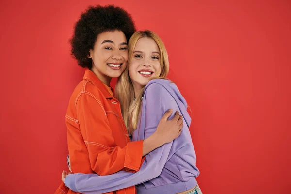 Cultural diversity, cheerful multicultural women hugging while looking at camera on coral background, blonde and brunette, diverse friends, sisterhood, friendship goals, studio shot — Stock Photo