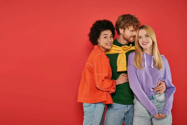 Polyamorous relationship concept, polygamy lovers, young man and multicultural women hugging on coral background, studio shot, denim fashion, love triangle, bonding and acceptance — Stock Photo