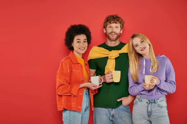 Open relationships concept, young multicultural women and redhead man holding cups on coral background, studio shot, vibrant colors, morning routine, coffee, happy polygamy lovers, polyamorous — Stock Photo