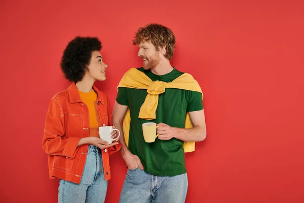 Multicultural couple holding cups of coffee on coral background, morning routine, cultural diversity, vibrant colors, stylish outfits, interracial lovers holding mugs and having conversation — Stock Photo