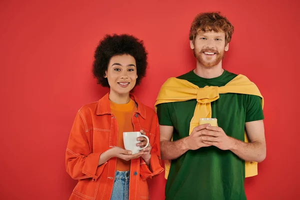 Multicultural couple holding cups of coffee on coral background, morning routine, cultural diversity, vibrant colors, stylish outfits, interracial people holding mugs and looking at camera — Stock Photo