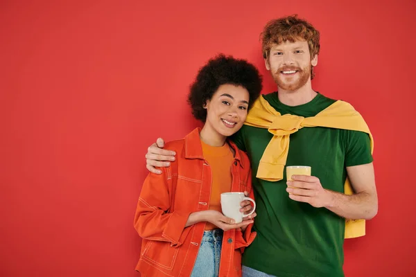 Multiracial couple holding cups of coffee on coral background, morning routine, cultural diversity, vibrant colors, stylish outfits, interracial people holding mugs and looking at camera — Stock Photo