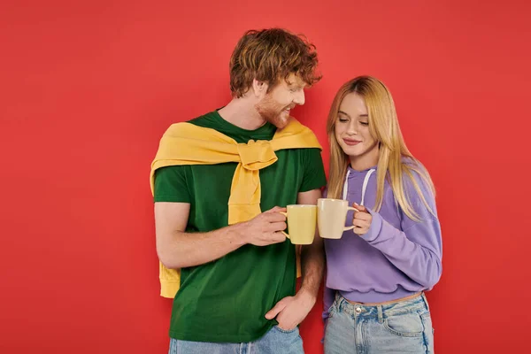 Happy young couple holding cups of coffee on coral background, morning routine, vibrant colors, stylish outfits, modern family, people holding mugs with hot drink, husband and wife — Stock Photo