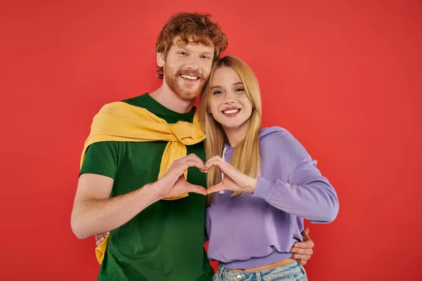 Portrait of young couple looking at camera on coral background, showing heart sign with hands vibrant colors, stylish outfits, modern family, husband and wife, bonding and love, togetherness — Stock Photo
