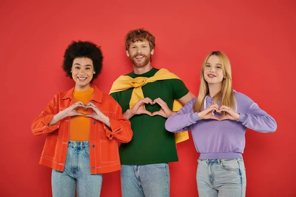 Open relationship, polygamy concept, three interracial lovers showing heart sign with hands on coral background, cultural diversity, polyamorous, happy multiethnic people looking at camera — Stock Photo