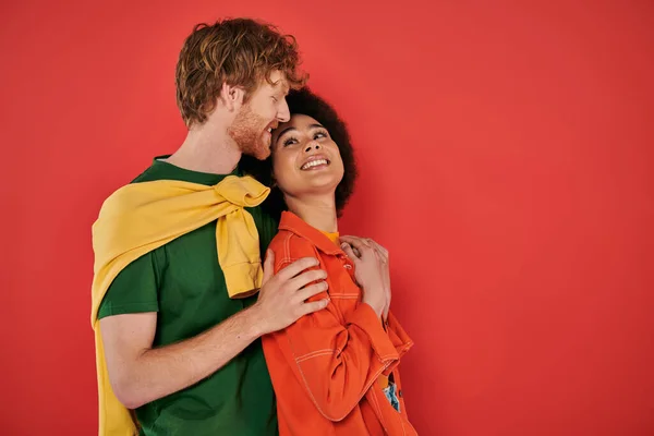 Interracial couple hugging and smiling on coral background, cultural diversity, vibrant colors, stylish outfits, youth and fashion, interracial people looking at each other, african american woman — Stock Photo