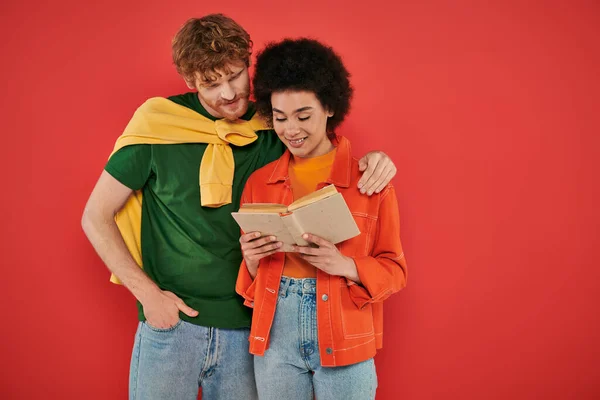 Interracial couple hugging and reading book on coral background, cultural diversity, vibrant colors, stylish outfits, youth and intelligence, multicultural people spending lovely time together — Stock Photo