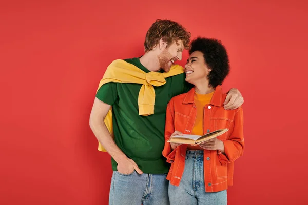 Interracial couple hugging and reading book on coral background, cultural diversity, vibrant colors, stylish outfit, youth and intelligence, multicultural people spending lovely time together — Stock Photo