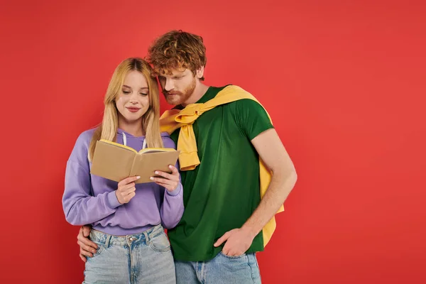Young couple hugging and reading book on coral background, vibrant colors, stylish outfit, youth and intelligence, blonde woman and redhead man spending lovely time together — Stock Photo