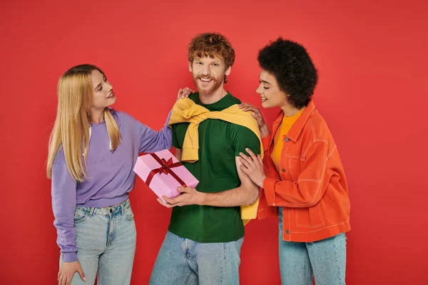 Open relationship, happy redhead man holding gift box near interracial bisexual women, polyamorous lovers smiling on coral background, holiday, festive occasions, alternative family — Stock Photo
