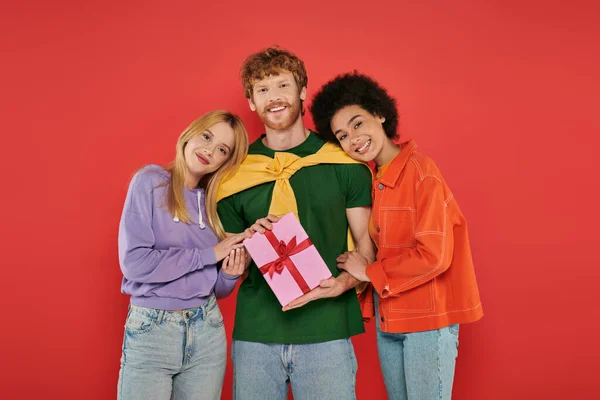Open relationship, polygamy, cheerful redhead man holding gift box near interracial bisexual women, polyamorous lovers smiling on coral background, holiday, festive occasions, alternative family — Stock Photo