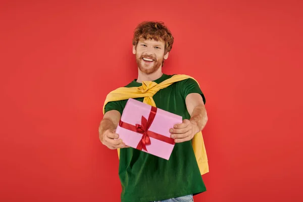 Holiday, cheerful redhead man with beard posing in casual attire on coral background, holding gift box, festive occasions, wrapped present, fashion and trend, urban style, happiness — Stock Photo