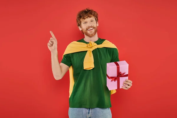 Празднование, счастливый рыжий мужчина с бородой позирует в повседневной одежде на коралловом фоне, держа подарочную коробку, праздничные мероприятия, подарок, мода и тенденция, счастье, праздник, указывая пальцем — стоковое фото