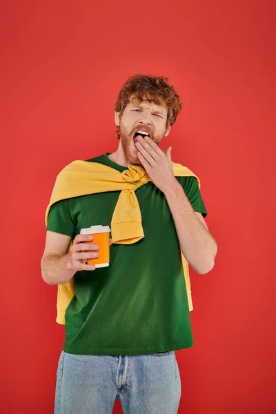 Morning coffee, redhead man with beard and curly hair holding paper cup on coral background, vibrant colors, male fashion, takeaway drink, sleepy man yawning and covering mouth — Stock Photo